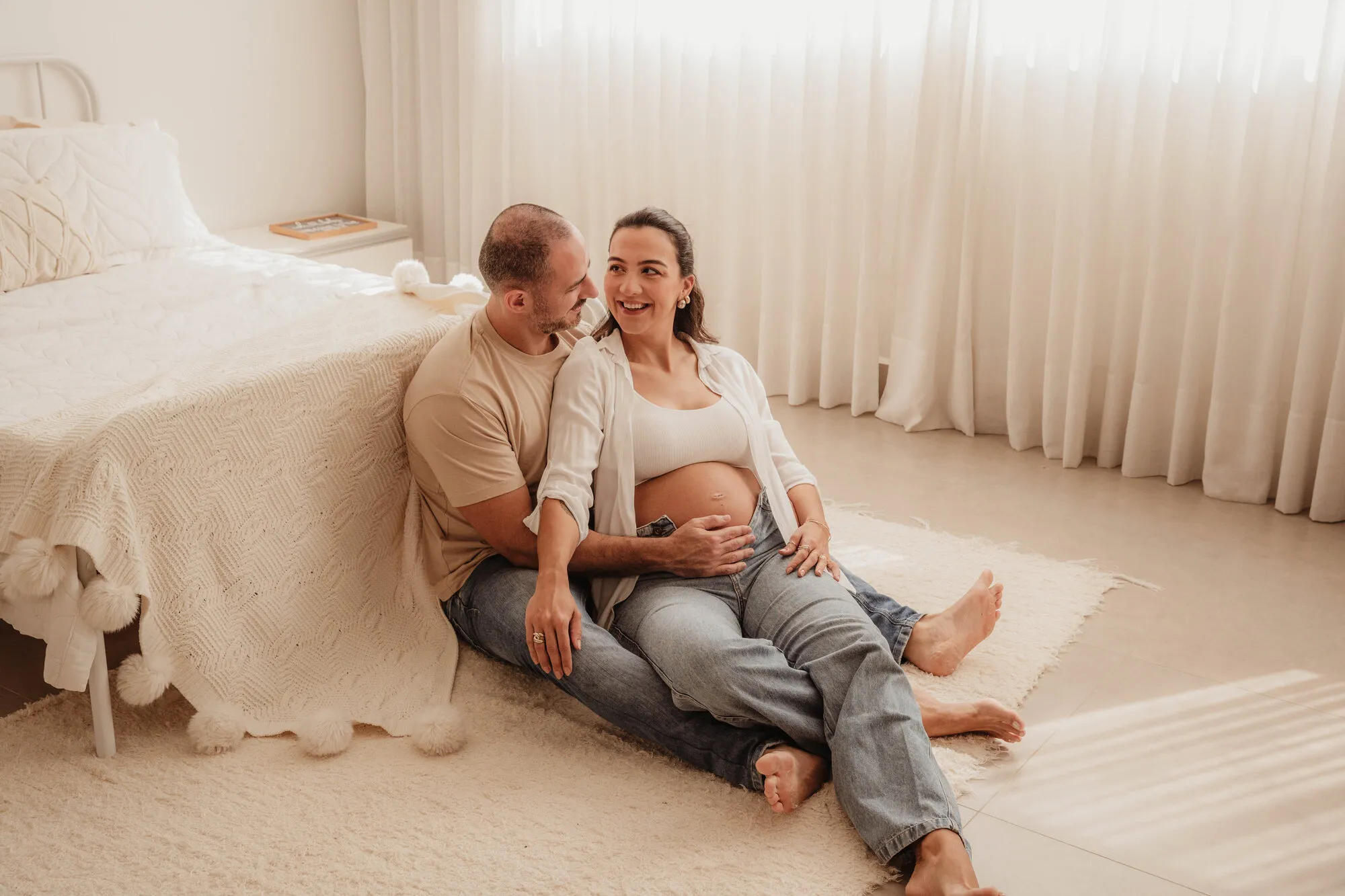 Casal grávido sentado no chão do quarto em ensaio de luz natural